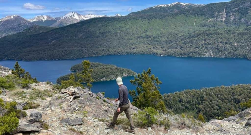 A person moves over rock terrain high above a blue body of water. In the distance there are snow capped mountains. 